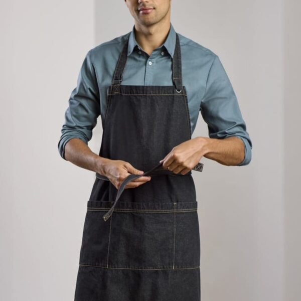 A person wearing a blue shirt and a black apron stands in a room, tying the apron strings. The room has neutral-colored walls and a wooden floor, giving it a simple and clean appearance.