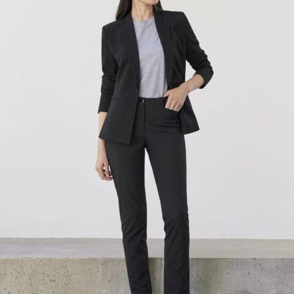 A woman stands against a plain background wearing a black blazer, black pants, and a gray top. She is also wearing black slip-on shoes. Her hands are relaxed by her sides, and she gazes to the right.