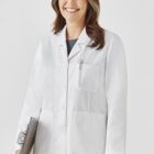Smiling person in a white lab coat holding a clipboard against a plain background.