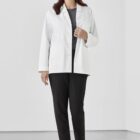 Person in a white coat and black pants smiling, standing in a light-colored room.