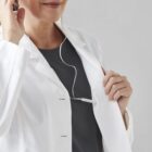 Person in a white coat adjusts white earbuds against a gray background.