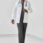 Smiling doctor in white coat and stethoscope stands in a bright room.