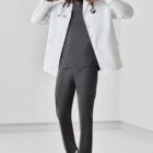 Person in a white coat with a stethoscope, wearing dark scrubs and sneakers, in a minimal room.