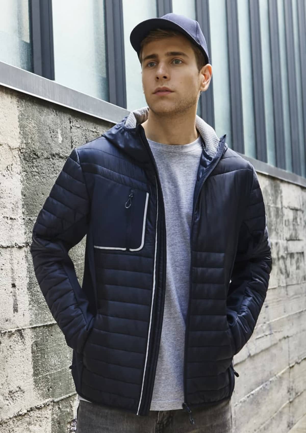 Young man in a navy jacket and cap stands near a concrete wall with windows behind him.