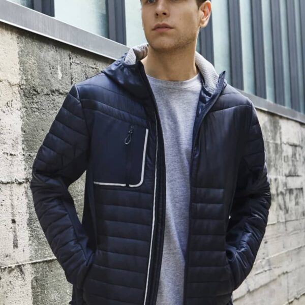 Man in a navy jacket and cap stands by a concrete wall with hands in his pockets.