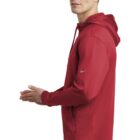 Man in a red hoodie standing sideways with hands clasped, looking slightly to the side.