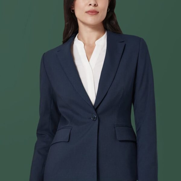 A person with long dark hair is wearing a navy blue suit and a white blouse. They are standing against a solid green background, looking straight ahead with a neutral expression.