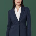 A person with long dark hair wears a navy blue suit jacket over a white blouse, standing against a solid green background. Their expression is calm and confident.