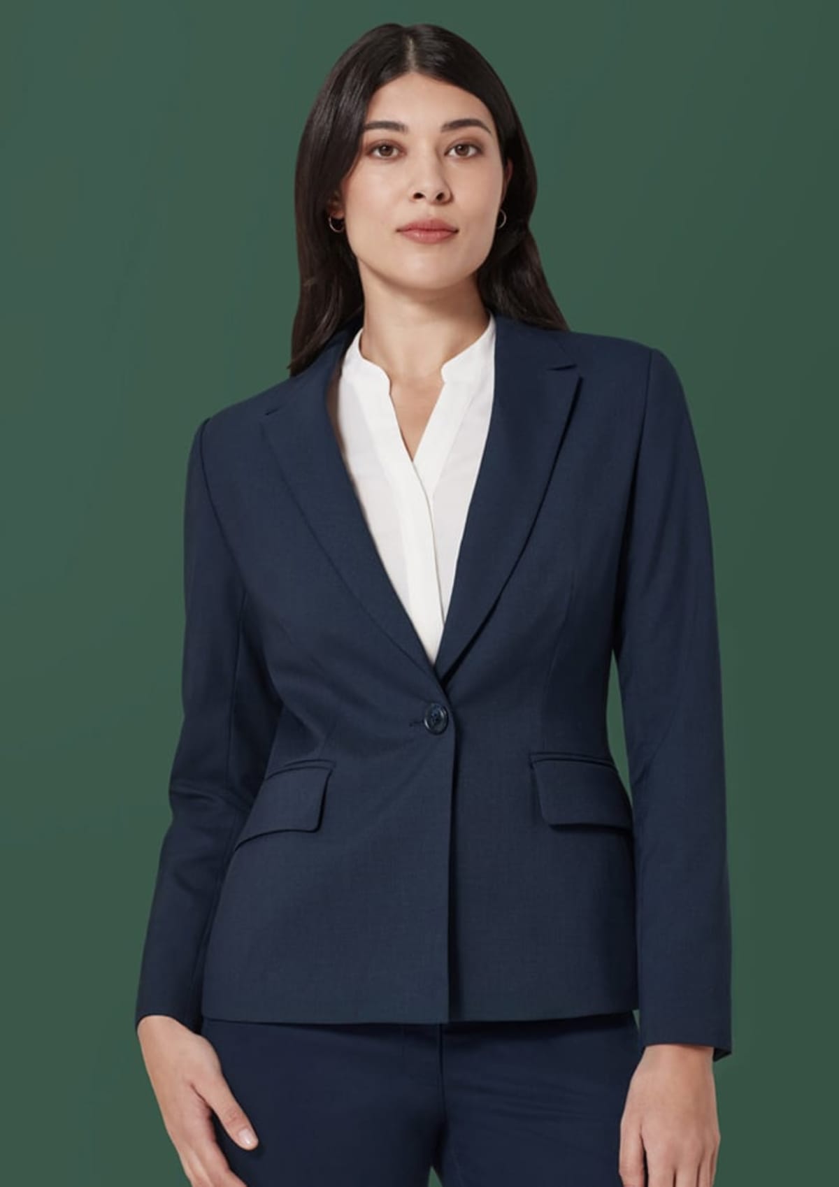 A person with long dark hair wears a navy blue suit jacket over a white blouse, standing against a solid green background. Their expression is calm and confident.