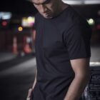 Person in a black shirt and beige pants stands pensively in a dimly lit urban setting.