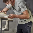 Person wearing a mask and working on a surfboard in a workshop.