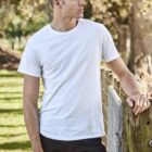 Man in a white t-shirt and black pants leans on a fence in a sunny outdoor setting.
