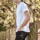 Person with curly hair in white shirt and jeans stands by a fence, gazing outdoors.