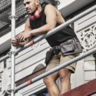 Worker in a black tank top, shorts, and tool belt standing on scaffolding, looking out.