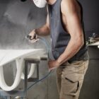 Person sanding a surfboard, wearing a mask, tank top, shorts, and cap.