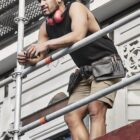 Man in shorts and tank top stands on scaffolding, wearing headphones and tool belt.