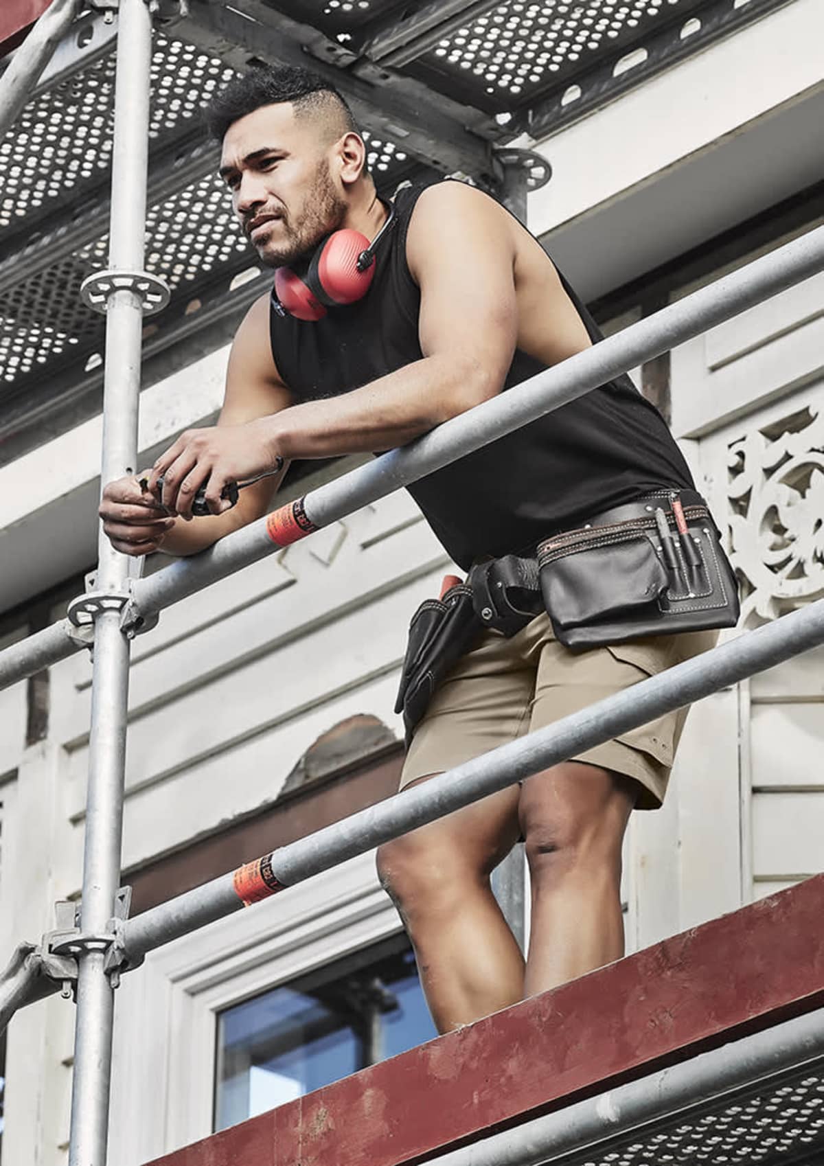 Man in shorts and tank top stands on scaffolding, wearing headphones and tool belt.