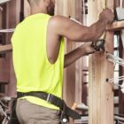 Man in a yellow tank top measures wooden beams with a tape measure at a construction site.