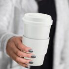 Hand holding a white reusable coffee cup with gray painted nails visible.