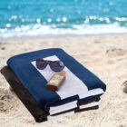Beach scene with sand and ocean in the background. In the foreground, three folded towels are stacked with a pair of sunglasses and a bottle of sun lotion on top. The towels are blue and white.