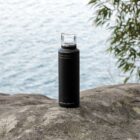 A black water bottle with a transparent lid is placed on a large rock. In the background, there is a view of water and some foliage on the right side.