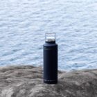 A black Arctic Zone branded water bottle with a clear cap sits on a rocky surface. The background features a view of the ocean with gentle waves.
