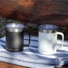 Two insulated mugs with clear lids are on a striped fabric. One mug is black, the other is white, both displaying the Arctic Zone logo. They are set on a wooden surface in an outdoor setting.