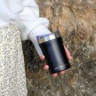 A person wearing a white sweater and floral skirt holds a black Arctic Zone tumbler with a transparent lid against a textured stone background.