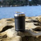 A black Arctic Zone tumbler with a clear lid sits on a sunlit rocky surface near a body of water, with blurred greenery and boats in the distant background.
