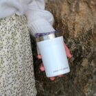 A person in a floral skirt and white sweater holds a white Arctic Zone tumbler with a clear lid. The background is a textured rock surface.