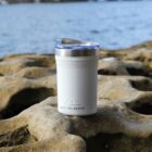 A white Arctic Zone tumbler with a clear lid is placed on a rocky surface by the water. The background features a blurred view of the ocean or a large body of water.