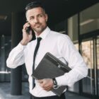 A man in a white dress shirt and black tie is holding a smartphone to his ear and carrying a black portfolio under his arm. He is standing outside a building, looking away from the camera with a slight smile.