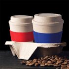 Two disposable coffee cups with red and blue sleeves sit in a cardboard holder on a dark surface, surrounded by scattered coffee beans. The background is black, highlighting the cups and beans.