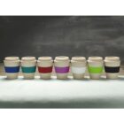 Seven reusable coffee cups lined up against a dark background. Each cup has a unique colored band: blue, green, red, pink, purple, white, and black. The cups are placed on a light-colored surface.