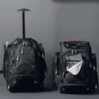 Two large black wheeled travel bags are shown against a gray background. One bag is upright with an extended handle, and the other is slightly open, revealing a laptop and headphones inside. Both bags have multiple zippers and red accents.