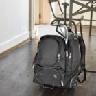 A black rolling backpack with multiple zippered compartments is placed on a wooden floor near a table and chair. It has a retractable handle and is partially unzipped, showing a white object, possibly a laptop, inside.