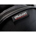Close-up of a black fabric bag featuring a black label with the word elleven in white lowercase letters, underlined with a thin red line. The texture of the fabric is visible, accentuated by lighting.