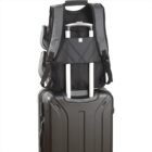 A gray backpack is securely placed on the handle of a black suitcase. The backpack is attached by a strap designed for easy transport on rolling luggage. The suitcase has a textured surface and a sturdy handle.