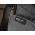 Close-up of a gray backpack with a black label displaying the word elleven in white text. The backpack features a zippered pocket and is set against a brick wall background.