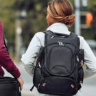 A person with brown hair tied in a bun is wearing a light jacket and carrying a large black backpack. They are walking alongside another person holding a suitcase. The background is blurry, showing greenery and street elements.