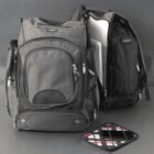 Two black and gray backpacks with multiple compartments sit on a gray surface. One backpack is open, revealing a laptop inside. In front of the backpacks is a small, plaid-patterned case.