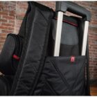 Close-up of a black backpack with a quilted texture, featuring a back sleeve that fits over a suitcase handle. The setting is a brick wall background, creating a travel-friendly vibe. Red accents are visible on the backpack straps.