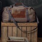 A gray duffel bag with brown leather accents and a front zipper pocket rests on top of a wooden crate. The crate has a metal handle leaning against it. The background is dark with indistinct writing.