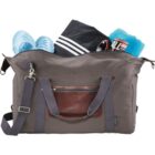 A gray duffel bag with brown leather accents is open, revealing its contents. Inside are light blue shoes, a black and white striped garment, and a rolled-up blue towel. A detachable shoulder strap is attached to the bag.