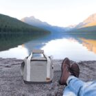 A pair of legs in blue jeans and brown boots rest on a rocky lake shore. A gray duffel bag sits nearby. The calm lake reflects the surrounding mountains and clear sky, creating a peaceful outdoor scene.