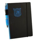 A black notebook with a blue spine and a school crest on the cover. Theres an elastic pen holder on the side holding a blue and silver pen.