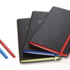 Three black notebooks with colored elastic bands (red, blue, and green) are stacked on a white surface. Two pens, one red and one blue, are placed beside the notebooks.
