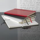 Stack of notebooks with red, black, and white covers on a reflective surface. An open notebook with lined pages is in the foreground. A blurred background shows handwritten cursive text.