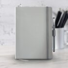 A gray notebook with a matching elastic band and attached pen sits on a white marble surface. The background is a blurred white brick wall and a white cup holding black pens.