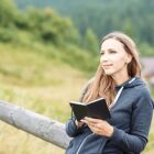 A woman with long hair is sitting outdoors near a wooden fence, holding an open notebook. She wears a dark hoodie and gazes into the distance with a slight smile. The background is a blurred green landscape.